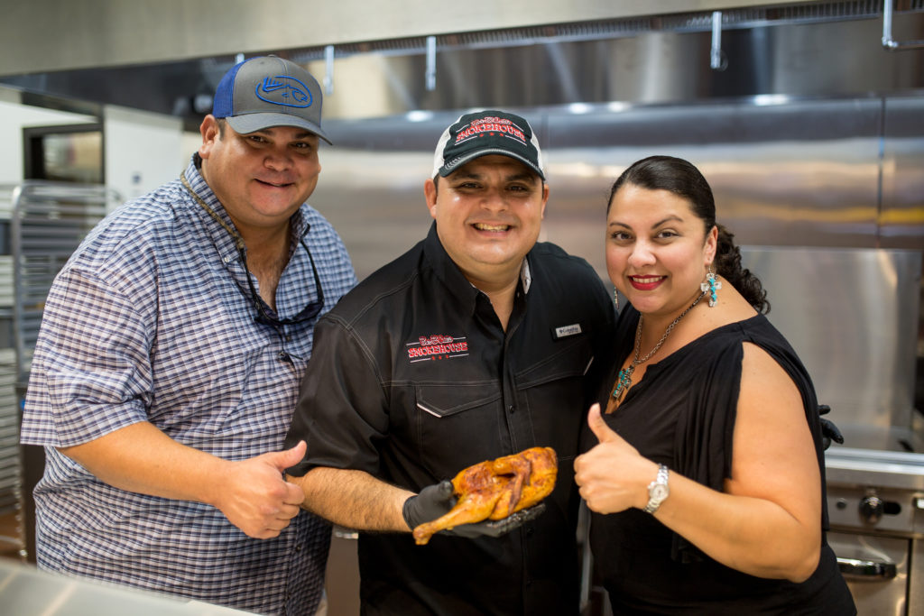 a smiling group of people at a Bar-B-Cutie kitchen holding a chicken leg and wing giving it a thumb's up